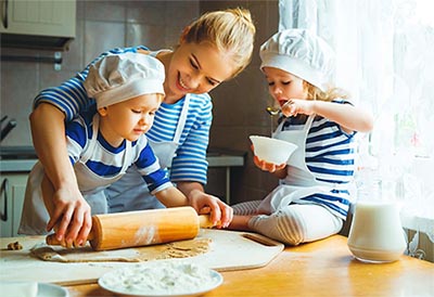 Una mamma prepara una torta con i suoi due bambini. Uno l'aiuta con il mattarello, mentre l'altra, con il cappello da chef, mangia dello yogurt