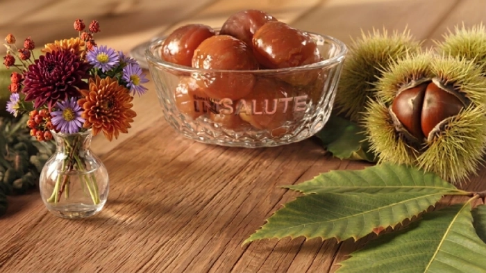 Tavolo con dei ricci e foglie di castagno con al centro una ciotola di cristallo con dentro dei Marron glacé. A sinistra dell'immagine è presente un vaso con dei fiori autunnali