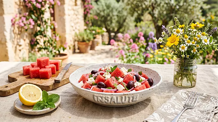 Su una tavola solare e colorata con un vaso di vetro con fiori di campo colorati, c'è un piatto, con su inciso "Obiettivo in Salute", con dell'insalata di anguria, olive nere, feta, menta, olio e limone. Sullo sfondo un giardino pieno di fiori e colori