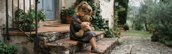 Una bambina seduta sulle scale esterne di una casa di periferia abbraccia, tristemente, il suo peluche