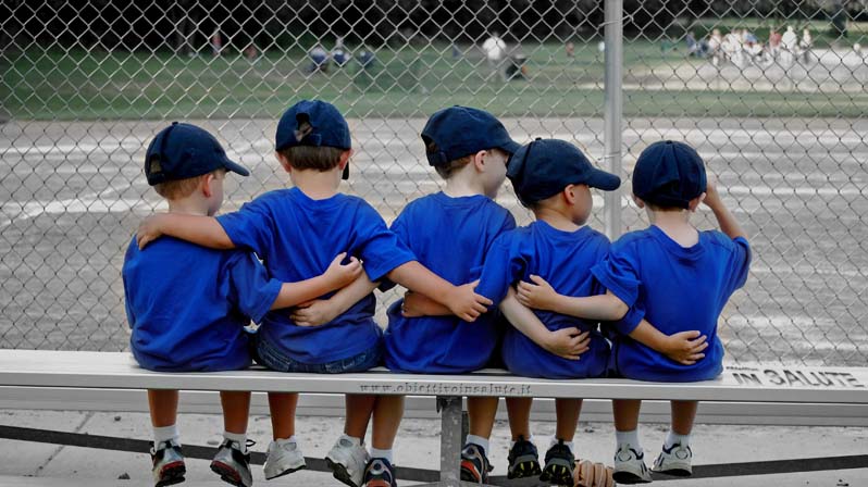 Cinque bambini seduti e abbracciati su di una panchina a bordo di un campo sportivo