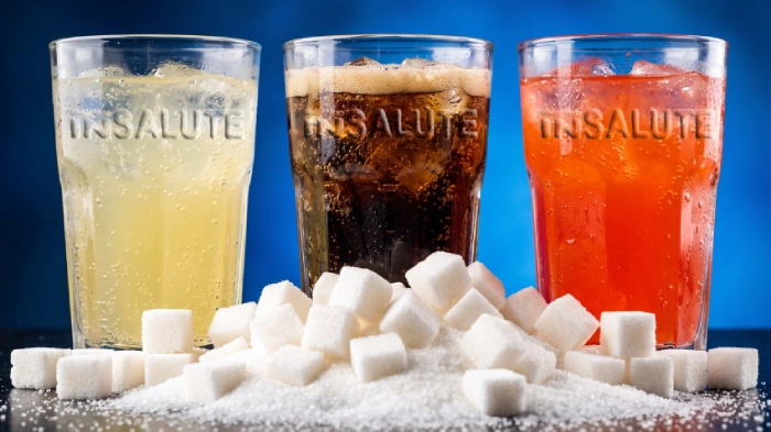 Tre bicchieri con bibite zuccherate di diverso colore, marrone, arancione e giallo, con davanti tanti cubetti di zucchero