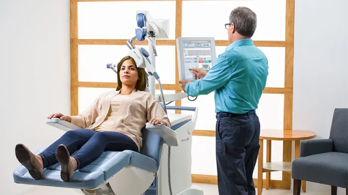 una donna è sdraiata sul lettino di uno studio medico con la testa poggiata su un macchinario che produce la stimolazione magnetica, mentre un dottore osserva i dati da uno schermo