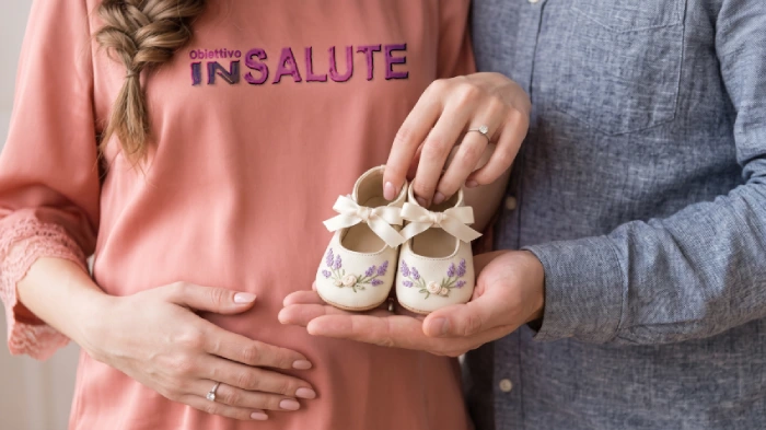Uomo è al fianco di una donna incinta e, tutti e due, tengono in mano delle scarpette da neonata