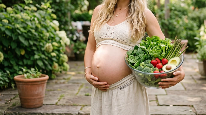 Donna in gravidanza, nel proprio cortile, ha in mano una ciotola trasparente con delle verdure e della frutta ricchi di acido folico come asparagi, lattuga, broccoli, spinaci, fragole e avocado